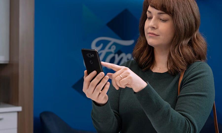 A red-haired woman uses her smartphone with a blue wall featuring the Ford logo in the background.