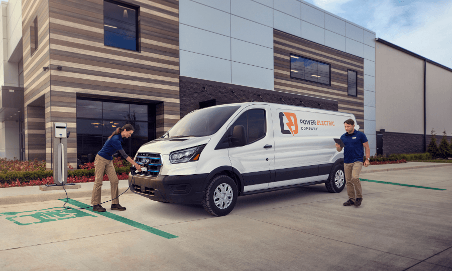 A female technician plugs a white Ford E-Transit electric van into a charging station while a male colleague looks at a tablet in front of a commercial building.