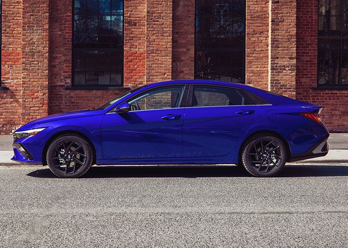 A red sedan is parked on a mountain road, with a large rocky cliff in the background.