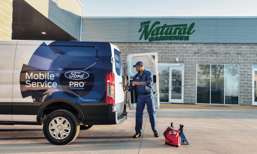 A mechanic in a blue uniform unloads tools from the back of a Ford Pro Mobile Service van parked in front of a business storefront.