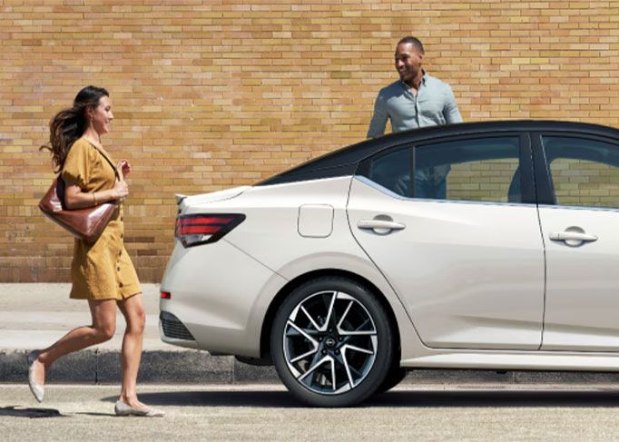 A red sedan is parked on a mountain road, with a large rocky cliff in the background.