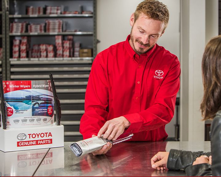 A Toyota parts advisor wearing a red Toyota shirt speaks with a customer at a parts counter. He is holding a product, possibly a windshield wiper blade, and pointing to it while talking to the customer. A display stand with a "TOYOTA GENUINE PARTS" sign is on the counter.