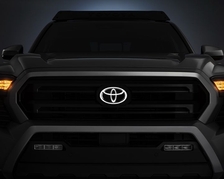 A low-light, head-on view of the front of a dark-colored Toyota truck. The grille is dark, and the headlights are turned on. The central Toyota emblem is illuminated, and the top of the windshield has a visible roof rack with "TOYOTA" spelled out in black.