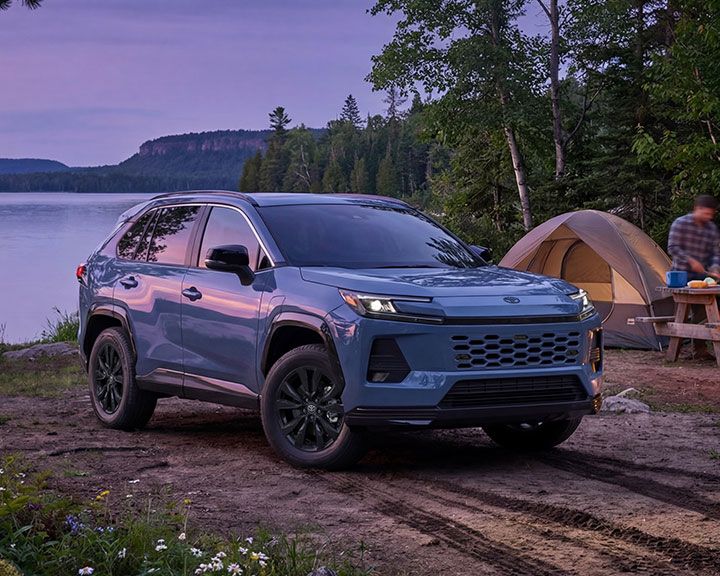 A smiling woman stands in a car dealership, looking at her smartphone while holding a takeaway coffee cup.