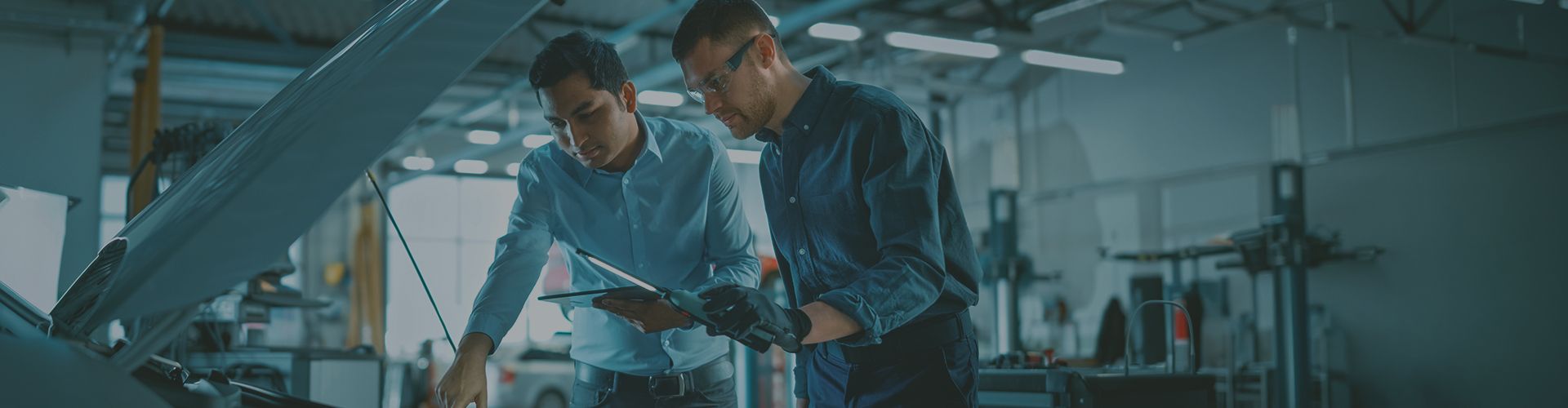 A mechanic with safety glasses and an inspector holding a tablet looking under the open hood of a car in an auto repair shop