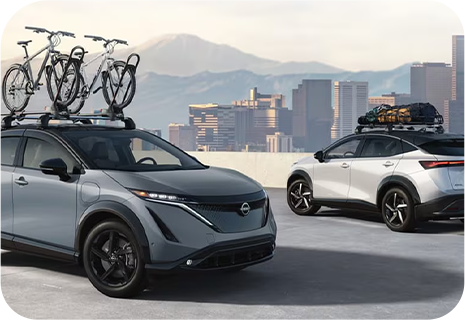 Two Nissan SUVs with roof racks carrying bikes and luggage overlooking a city skyline.