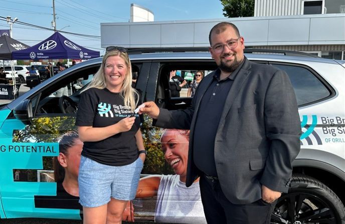 A man in a grey blazer hands a set of car keys to a smiling woman wearing a "Big Brothers Big Sisters of Orillia and District" t-shirt. They are standing next to a Volkswagen SUV wrapped with the organization's branding and a large photo of a smiling child.