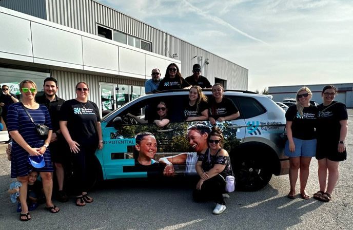 A large group of people poses for a photo around and inside a branded "Big Brothers Big Sisters" Volkswagen SUV. Most are wearing matching black t-shirts with the organization's logo, and the vehicle features a colorful wrap with the words "IGNITING POTENTIAL."
