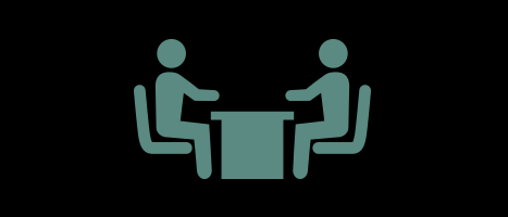 A sage-green minimalist icon on a black background depicting two people sitting across from each other at a table, symbolizing a meeting, conversation, or community connection.