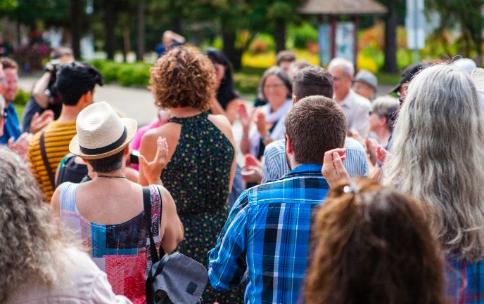 A view from behind a diverse crowd of people gathered outdoors in a park-like setting. Several individuals are clapping their hands, suggesting they are watching a performance or presentation.