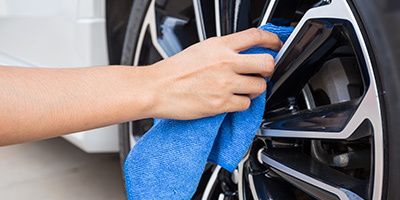 A close-up shot of a hand using a blue microfiber cloth to carefully clean the intricate silver and black rim of a car wheel.