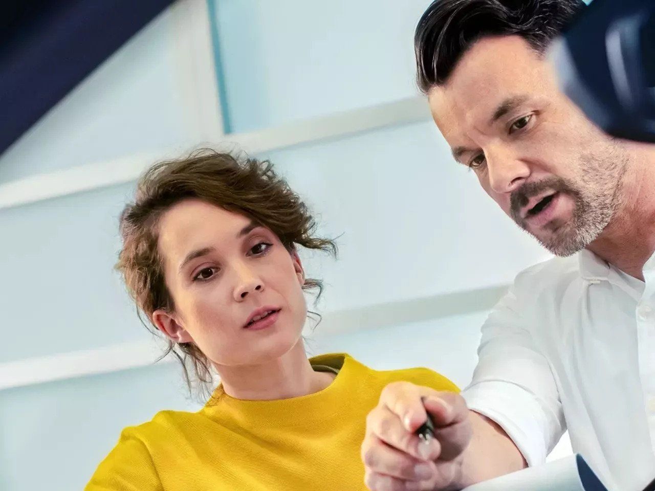 A low-angle, close-up shot of a woman in a yellow sweater and a man in a white shirt reviewing a document together; the man is pointing at a specific detail with a pen while both maintain a focused expression.