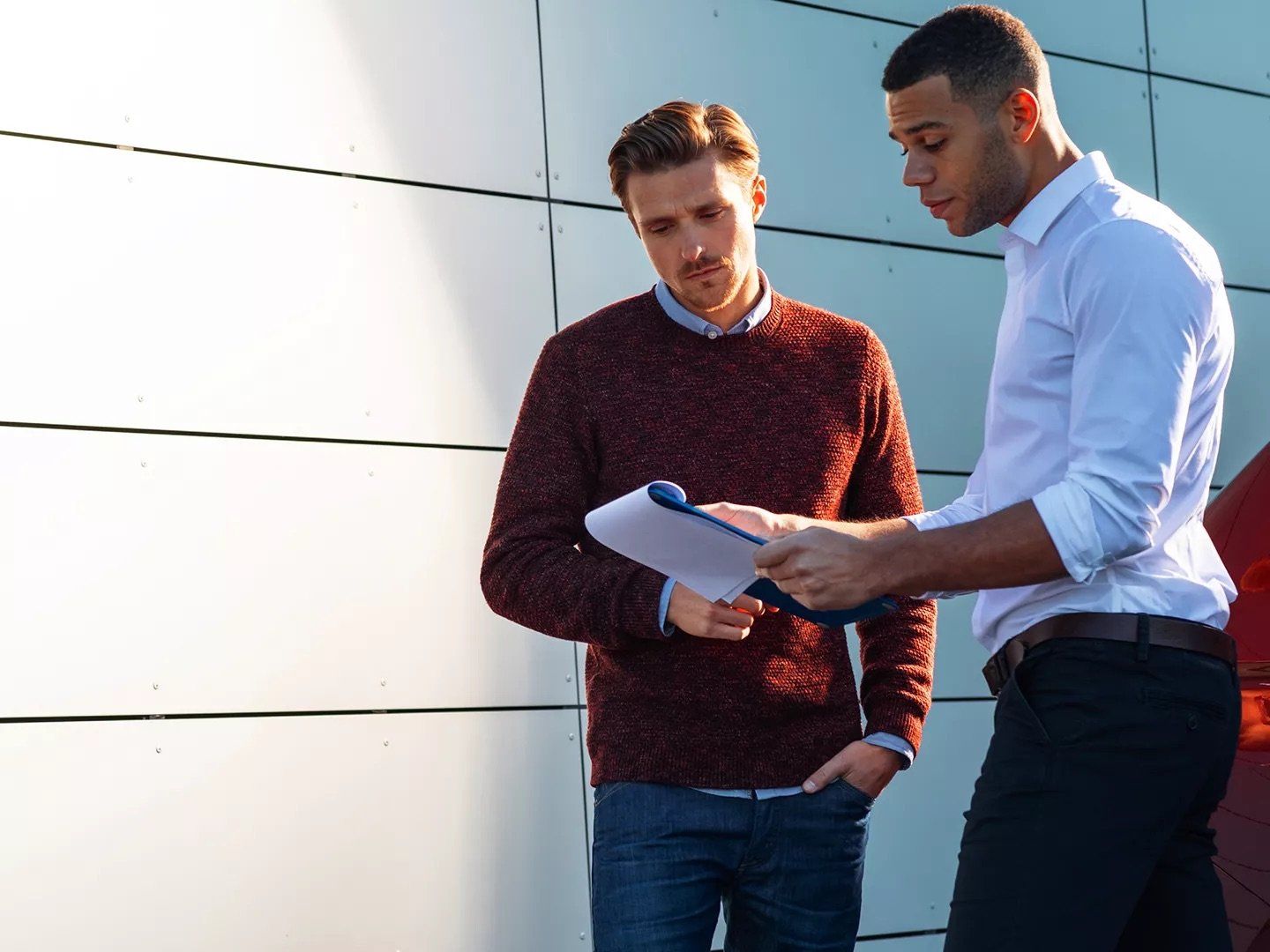 A man in a maroon sweater and a man in a white dress shirt stand outside next to a modern building, looking down at papers held in a blue clipboard.