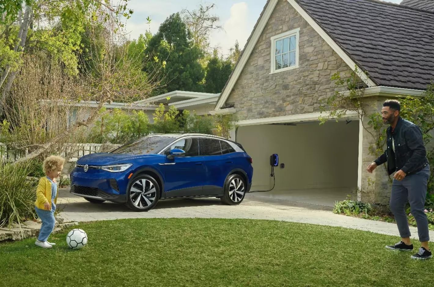 A blue VW ID.4 parked in a driveway while a man and a young child play soccer on the lawn nearby.
