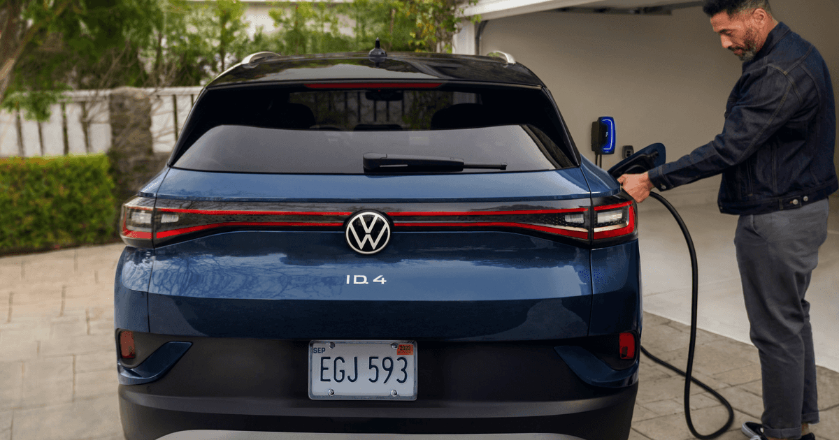 A man plugging a charging cable into the rear passenger-side port of a blue VW ID.4 in a driveway.