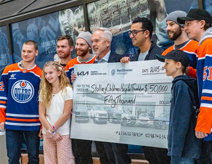 A group of people, including several men in Edmonton Oilers hockey jerseys and two children, stand together for a photo. In the center, two men hold a large ceremonial donation check for $50,000 made out to the Stollery Children's Hospital Foundation from North Edmonton Kia and West Edmonton Volkswagen.