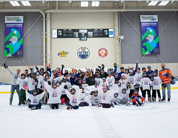 A large group of children and adults pose for a photo on the ice at the Downtown Community Arena. The children are wearing white "Skate for Cancer" jerseys, while several adults wear Edmonton Oilers jerseys. The arena scoreboard and team logos are visible in the background.