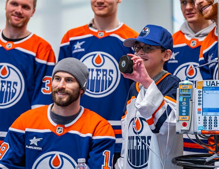 A young boy wearing an Edmonton Oilers jersey, glasses, and a medical oxygen tube smiles while holding up a hockey puck. He is surrounded by several smiling Edmonton Oilers players in team jerseys. A medical infusion pump is visible in the foreground.