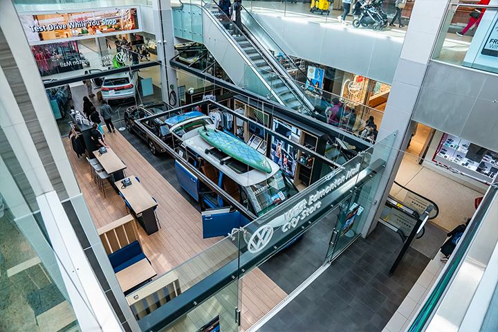 A high-angle shot looking down into the store, capturing the "Test Drive While You Shop" digital banner and the integration of the cars within the mall's architecture.