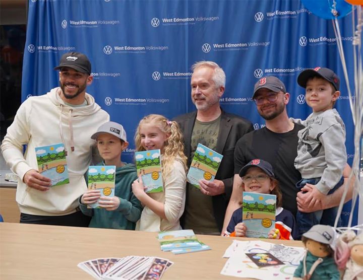 A group of children and men pose together holding copies of a children's book. The background is a blue step-and-repeat backdrop featuring the West Edmonton Volkswagen logo.