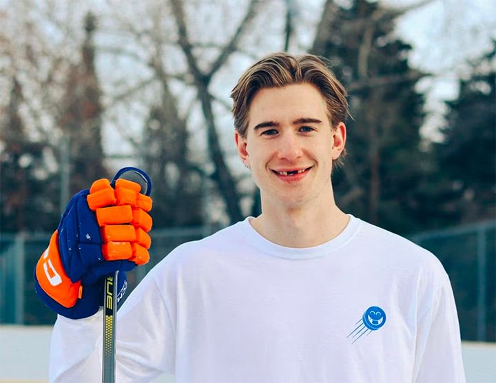A close-up portrait of a young man smiling at the camera. He is wearing a white long-sleeve shirt with a blue puck logo and holding a hockey stick. He has a visible gap in his front teeth, typical of a hockey player.