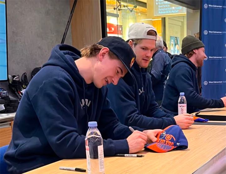 Two men in dark hoodies sit at a wooden table during an autograph session. The man in the foreground is signing an orange and blue Edmonton Oilers hat with a permanent marker.