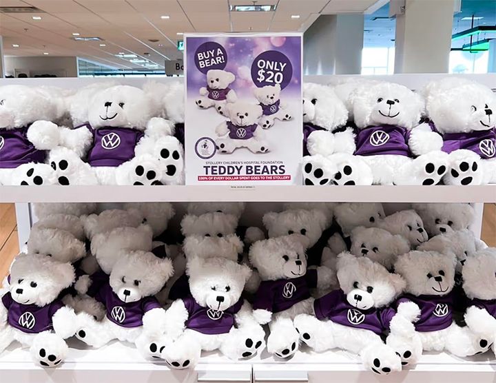 Shelves filled with white teddy bears wearing purple sweaters with the Volkswagen logo. A sign in the center advertises the bears for $20, noting that 100% of proceeds go to the Stollery Children's Hospital Foundation.