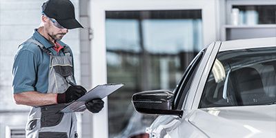 A mechanic in a grey uniform and black cap stands next to a white car, focused on writing notes on a clipboard.