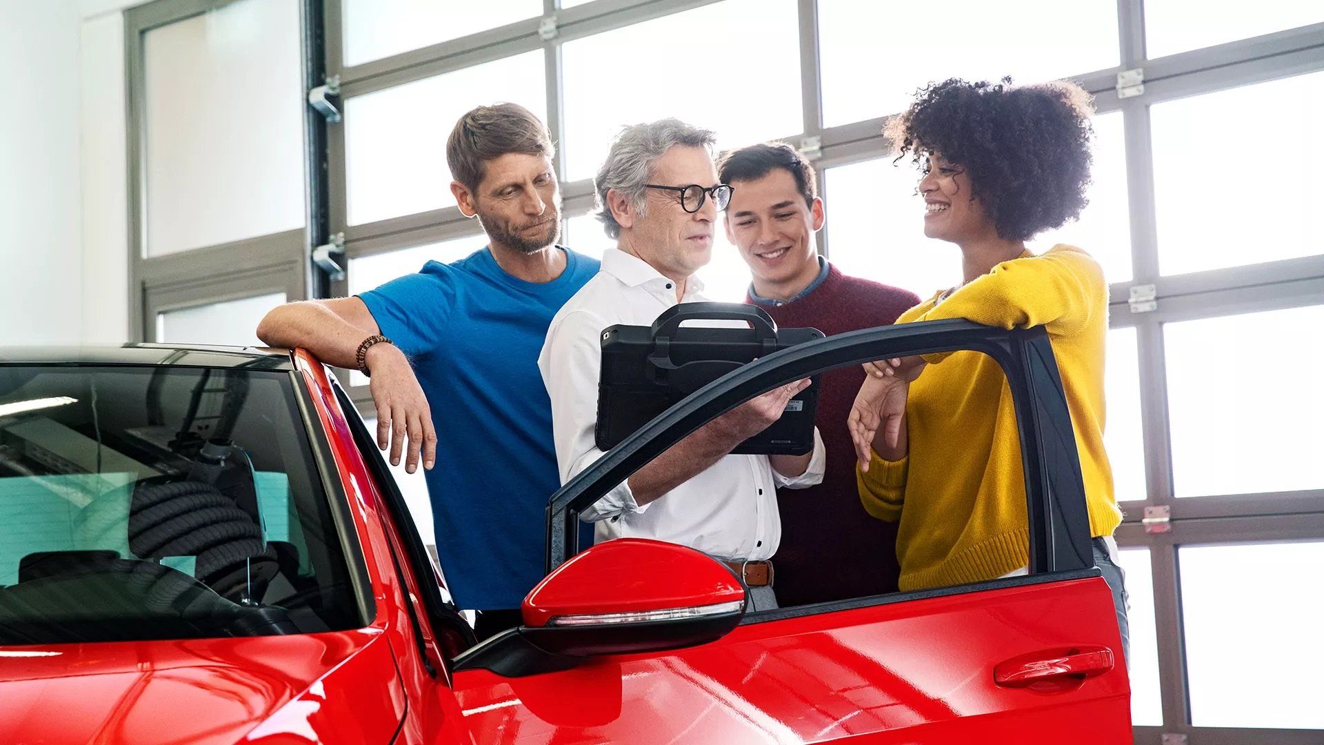 A group of four people—three men and one woman—gather around a red car inside a brightly lit showroom; a man in a white shirt holds a tablet while the others look on with interested and smiling expressions.