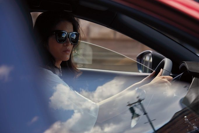A smiling woman stands in a car dealership, looking at her smartphone while holding a takeaway coffee cup.