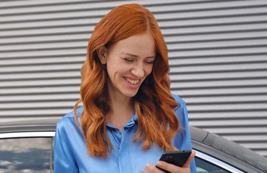 A smiling red-haired woman in a blue shirt looks down at her smartphone next to a car.