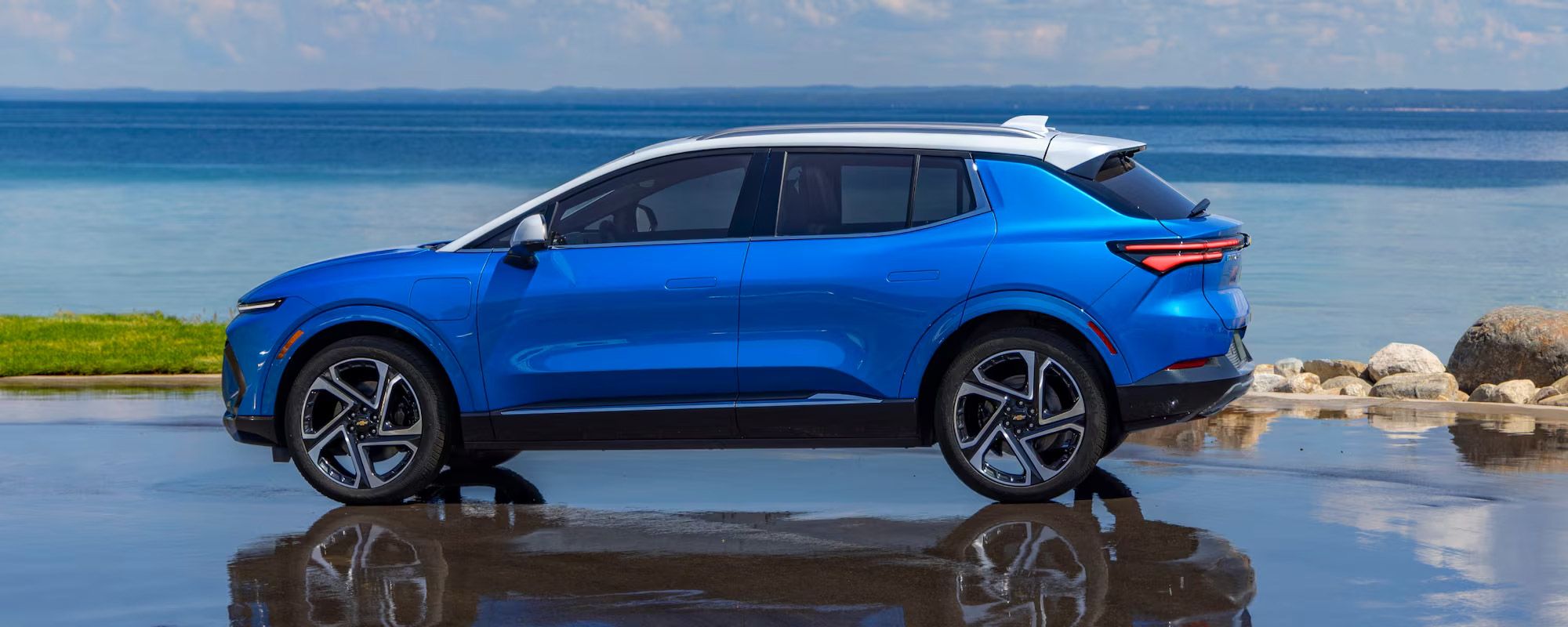 Une Chevrolet Equinox EV bleue avec un toit blanc est garée sur une surface mouillée, reflétant le véhicule, avec un grand plan d'eau et une rive rocheuse en arrière-plan sous un ciel nuageux.