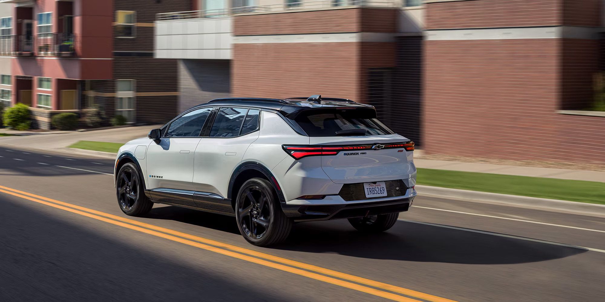 Une Chevrolet Equinox EV blanche avec un toit noir s'éloigne sur une route pavée, passant devant des bâtiments résidentiels modernes. Les feux arrière de la voiture sont allumés, et « EQUINOX EV » est visible à l'arrière.