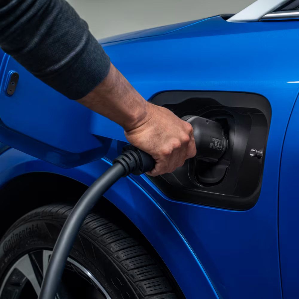 Une main est montrée en train de brancher un câble de recharge noir dans le port de recharge d'un véhicule électrique bleu, avec une partie du pneu avant visible en dessous.
