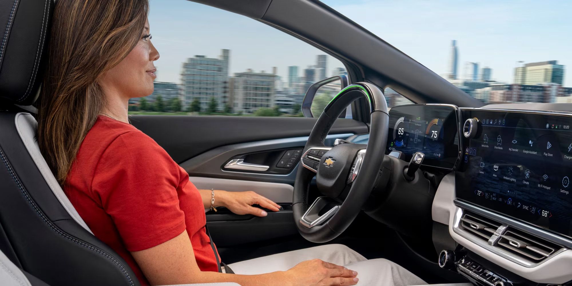 Une femme aux cheveux bruns et longs, vêtue d'un t-shirt rouge et d'un pantalon blanc, est assise sur le siège conducteur d'une Chevrolet Equinox EV. Elle regarde droit devant elle, et l'intérieur de la voiture, y compris le volant et les grands écrans numériques, est visible. À travers la fenêtre, un paysage urbain flou suggère que la voiture est en mouvement.