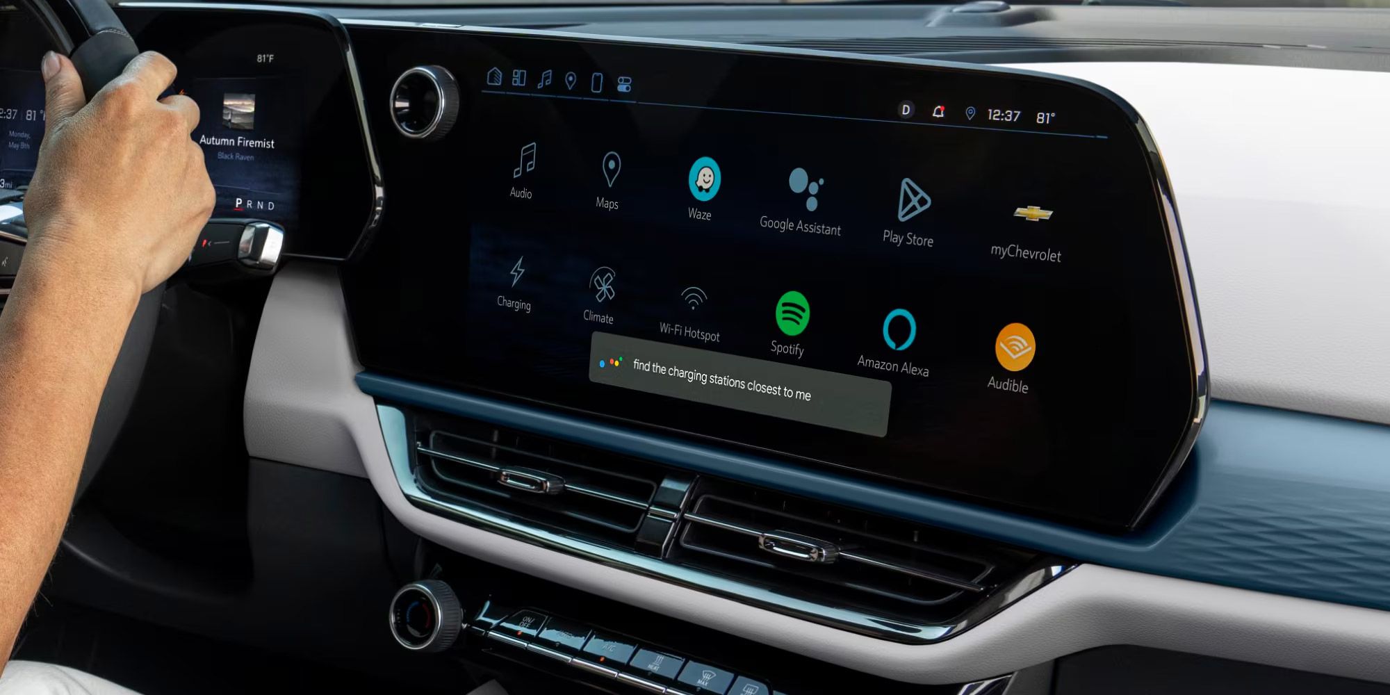 Vue rapprochée de l'écran d'infodivertissement à l'intérieur d'une Chevrolet Equinox EV. Le grand écran tactile affiche diverses icônes d'applications, notamment Audio, Cartes, Waze, Assistant Google, Play Store, myChevrolet, Recharge, Climatisation, Point d'accès Wi-Fi, Spotify, Amazon Alexa et Audible. Une invite sur l'écran indique « trouver les bornes de recharge les plus proches de moi ». Une main est visible sur le volant à gauche.