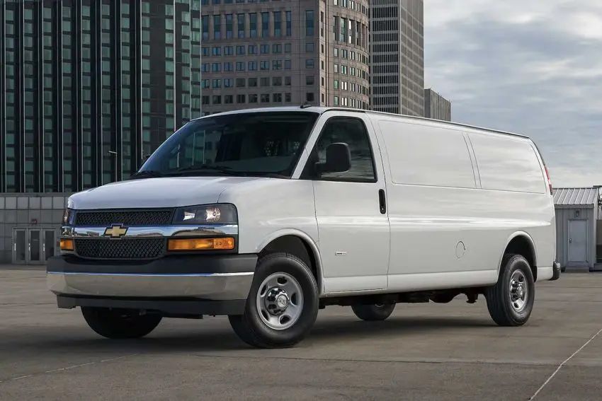 A white Chevrolet Express cargo van, shown from a low angle, parked on pavement in a city center or business district on an overcast day.