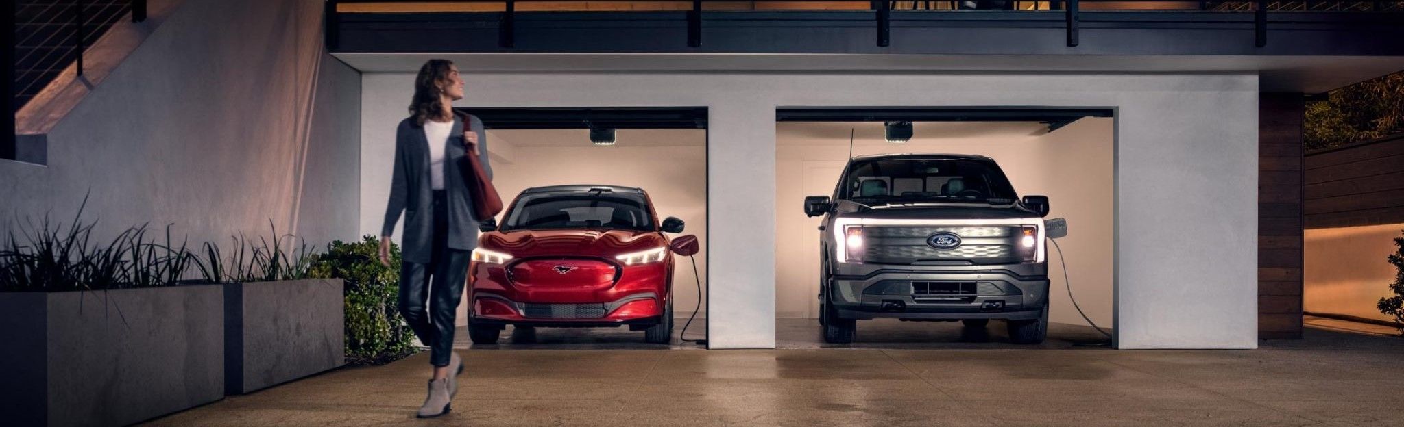 Woman walking out from home garage with a 2024 Mustang® Mach-E and Ford Lightning® truck in plugged-in and charging