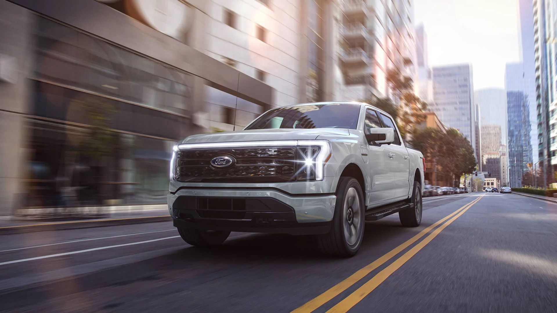 A light gray Ford F-150 Lightning electric pickup truck drives on a city street, with modern buildings and other vehicles visible in the background.