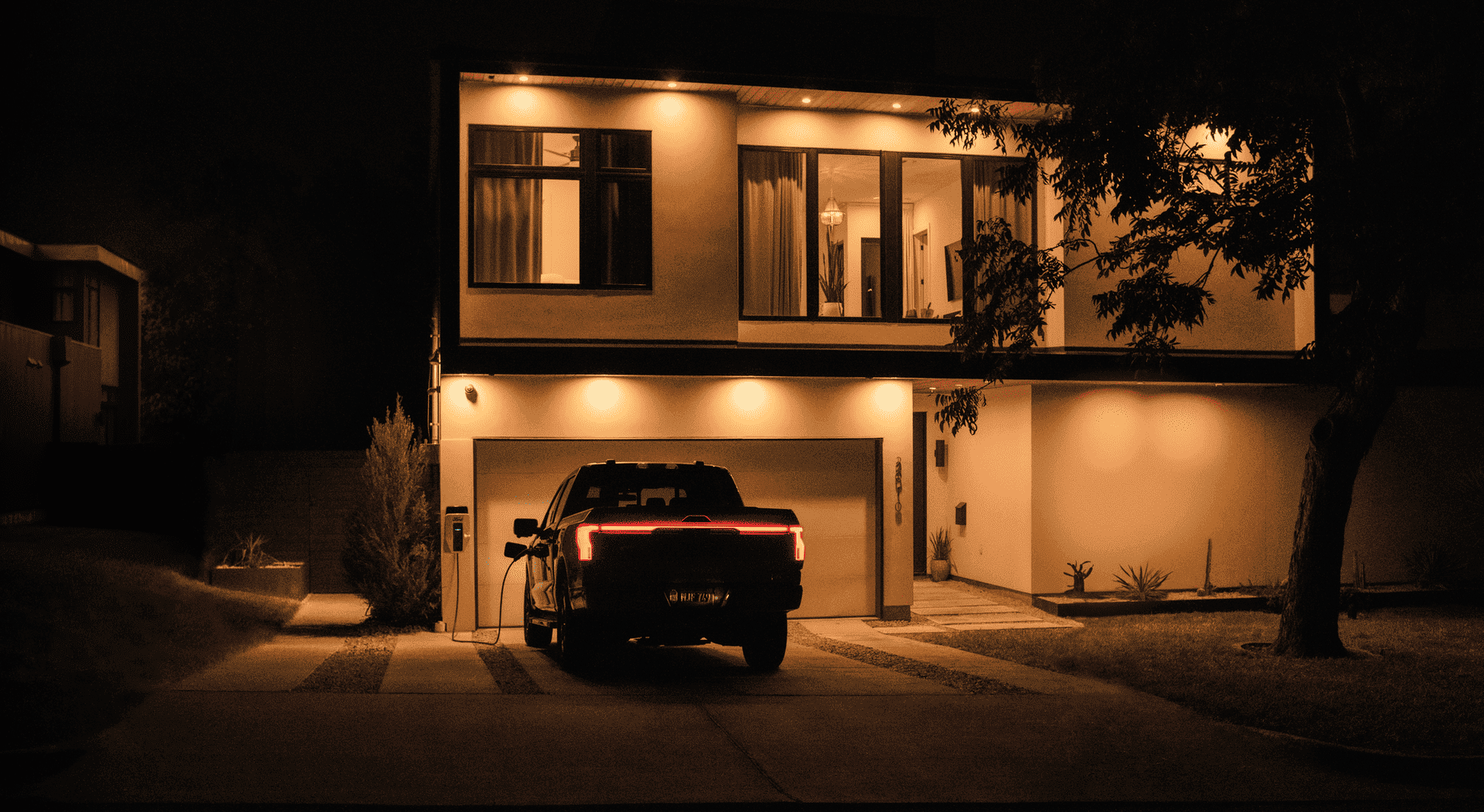 A dark-colored Ford F-150 Lightning electric pickup truck is parked in a driveway at night, plugged into a home charging station, with the rear lights illuminated and a modern house lit from within and by exterior spotlights in the background.