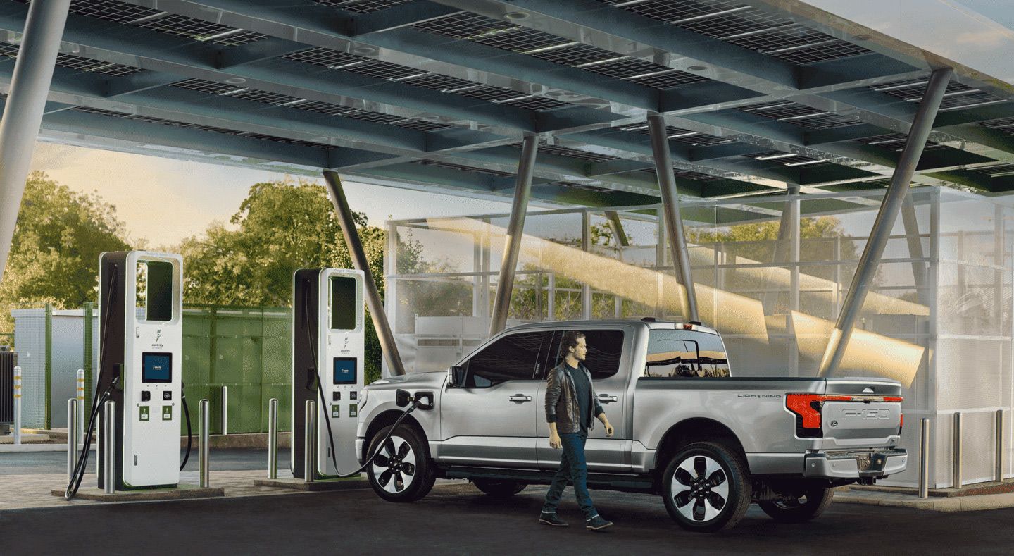 A silver Ford F-150 Lightning electric pickup truck is parked under a solar-panel covered charging station, with a man walking alongside it towards two charging pillars.