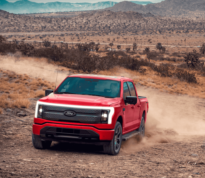  A red Ford F-150 Lightning electric pickup truck drives on a dirt road, kicking up dust, with a rocky, arid landscape and mountains in the background under a cloudy sky.