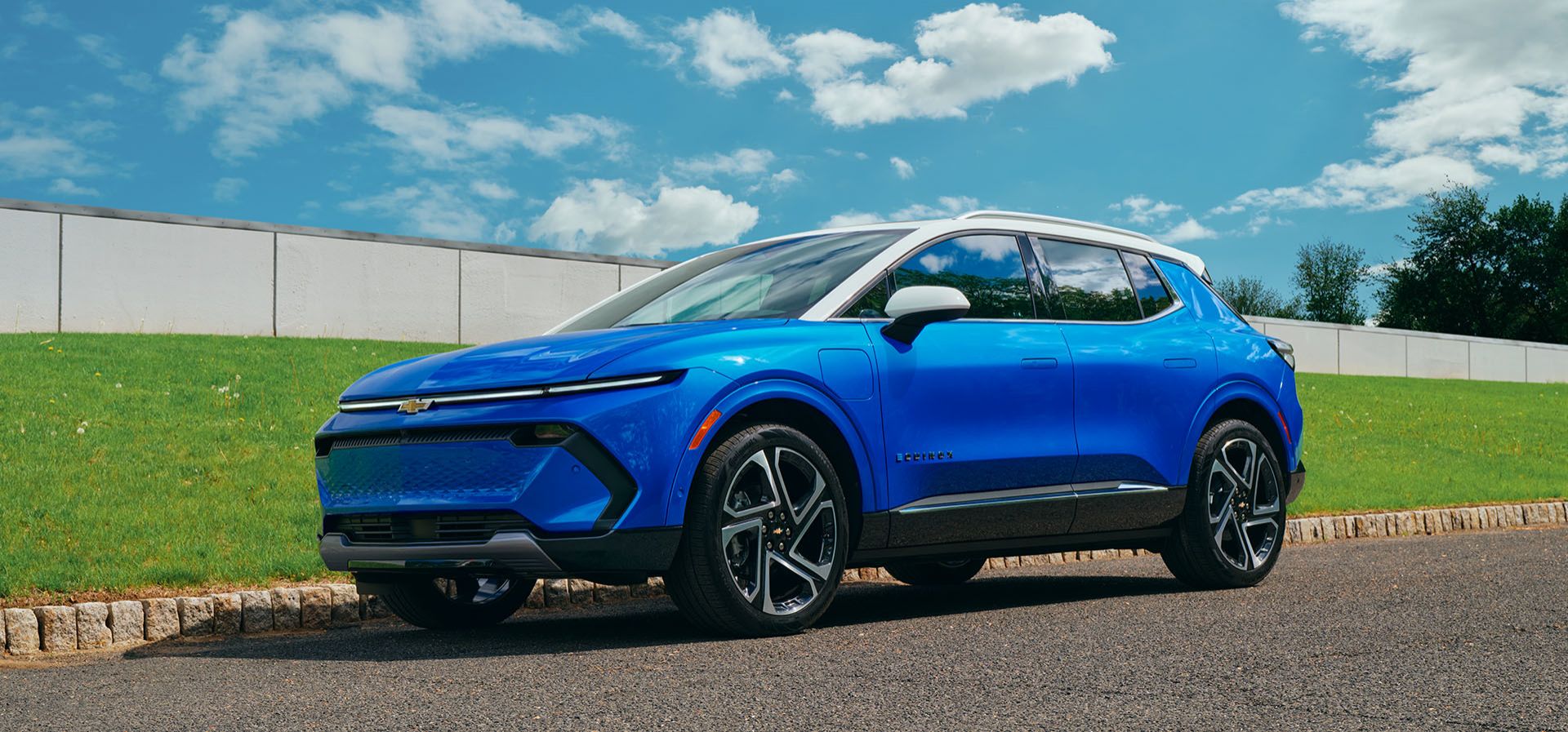 Une Chevrolet Equinox EV gris clair roule sur une route pavée à côté d'un plan d'eau avec des arbres et des collines en arrière-plan sous un ciel dégagé.