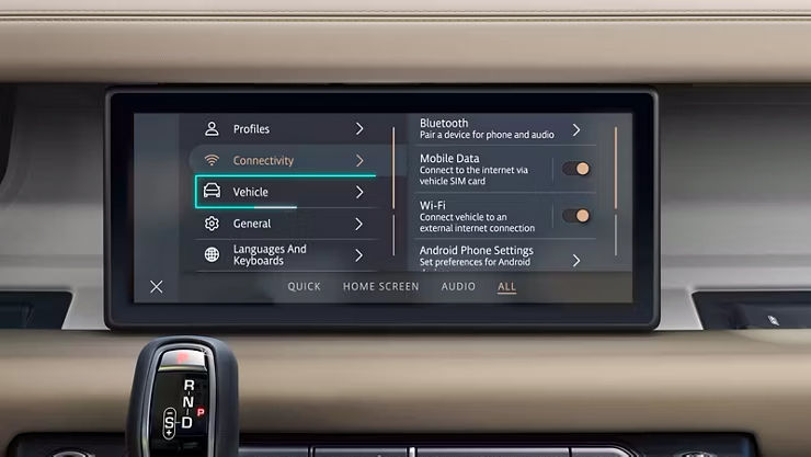 Close-up of a Land Rover infotainment touchscreen displaying the vehicle settings menu, with 'Connectivity' and 'Vehicle' options visible, alongside a gear shifter in the foreground.