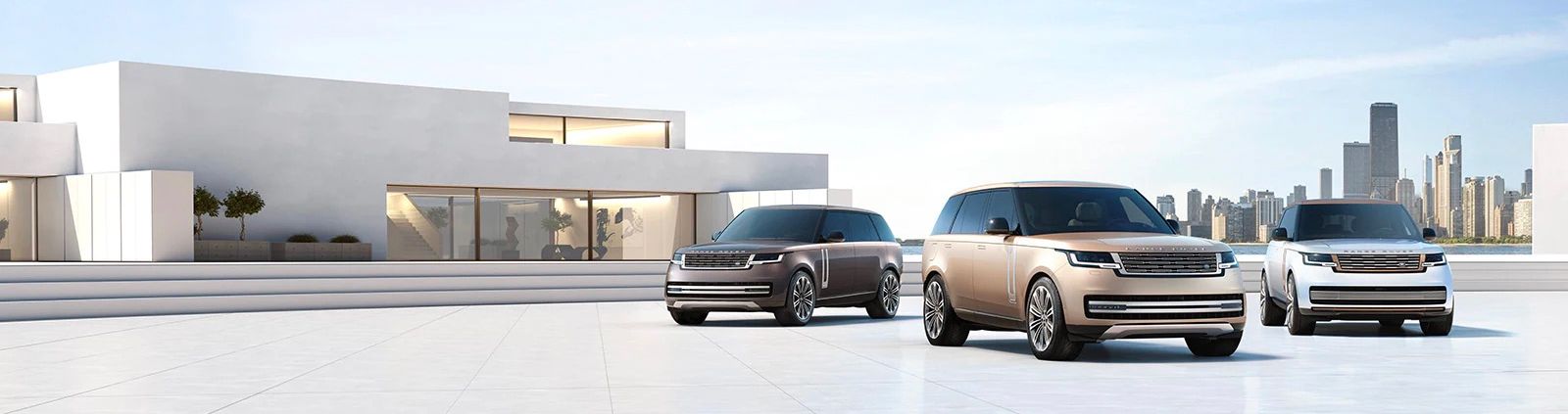 Three Land Rover SUVs – a dark grey, a champagne gold, and a white – parked in front of a modern, white dealership building with a city skyline in the background.