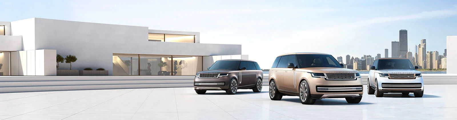 Three Land Rover SUVs – a dark grey, a champagne gold, and a white – parked in front of a modern, white dealership building with a city skyline in the background.