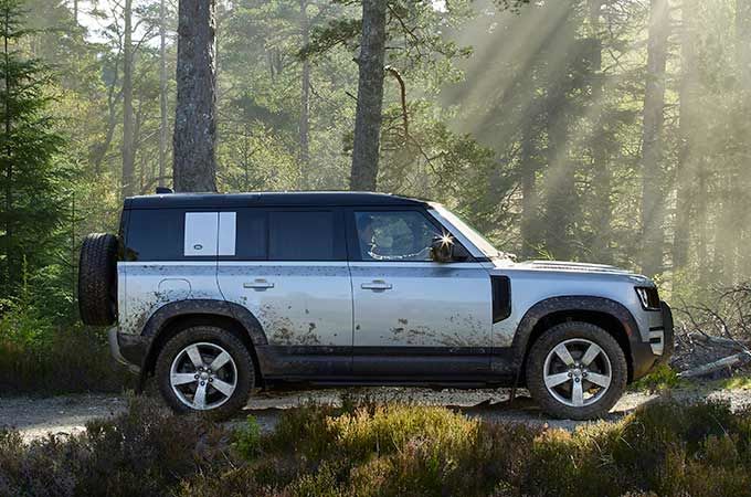 A silver Land Rover Defender with a black roof, lightly splattered with mud, parked on a dirt track in a sunlit forest.