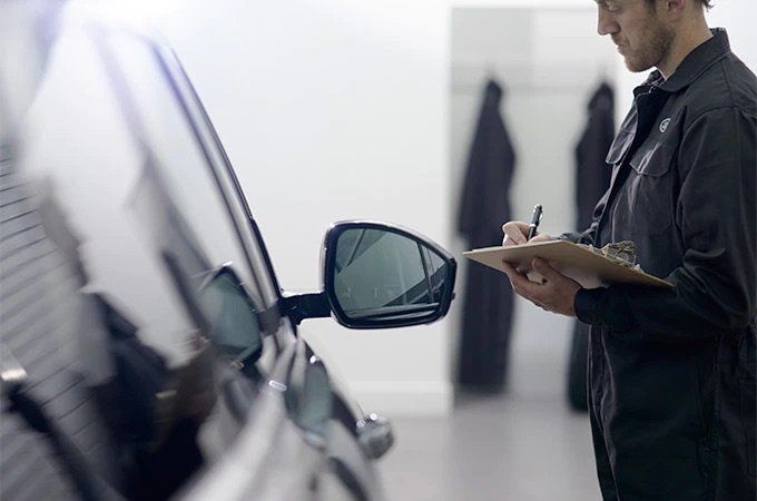 A Land Rover service technician in dark overalls inspects the side of a black vehicle while writing on a clipboard in a bright, clean workshop.