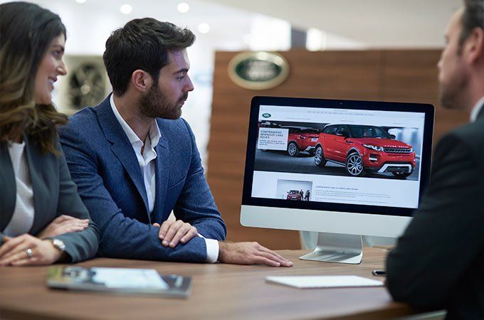 A couple sits at a desk in a Land Rover dealership, looking at a computer screen displaying a red Land Rover Evoque on the Land Rover website.