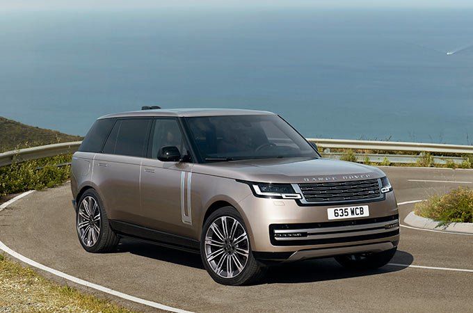 A champagne gold Range Rover driving along a winding coastal road with hills and the sea in the background.